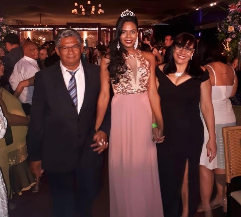 Foto de uma família. Uma mulher usando um vestido de festa rosa longo está de mãos dadas com um homem e uma mulher. A mulher ao lado direito usa um vestido preto. O homem do outro lado está de terno. Atrás deles, está uma festa com muitos convidados.